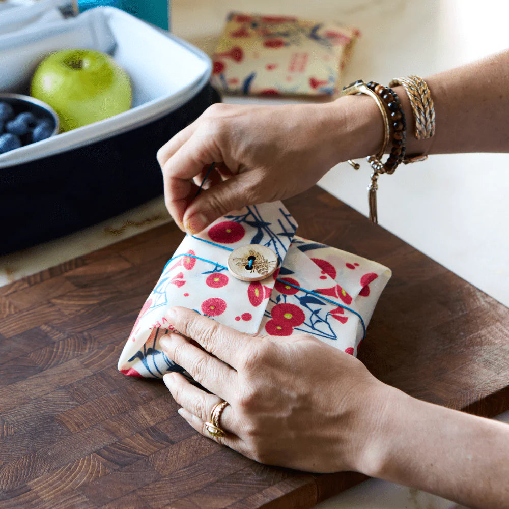 Hands folding and buttoning Bee's Wrap Lunch Pack Wrap - Full Bloom on a wooden board with snacks nearby.