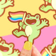 Rainbow Frog Sticker showing a happy green frog holding a waving rainbow pride flag on a colorful background.