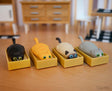 Racing Cat in Box toys in assorted colors, each featuring a cat snug in a yellow cardboard box on a wooden table.