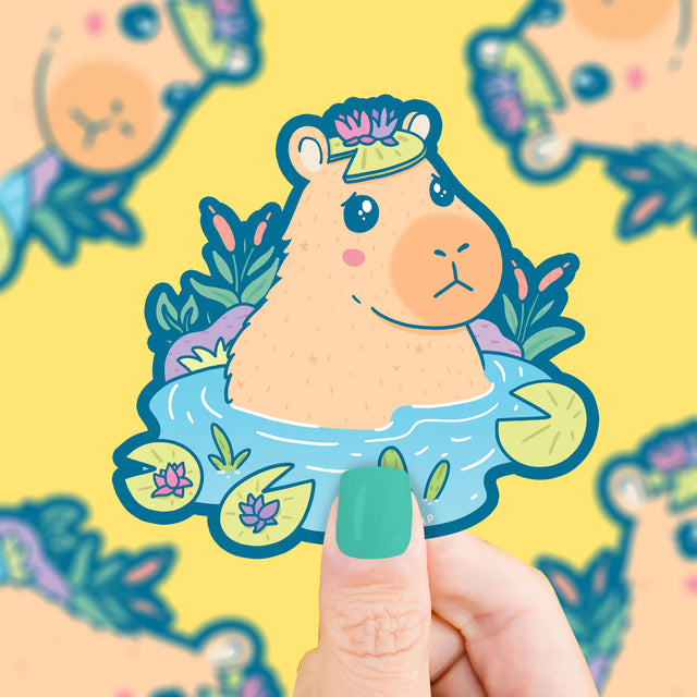Capybara Pond Sticker showing a cute capybara with a lily pad hat sitting in a pond with flowers and leaves.