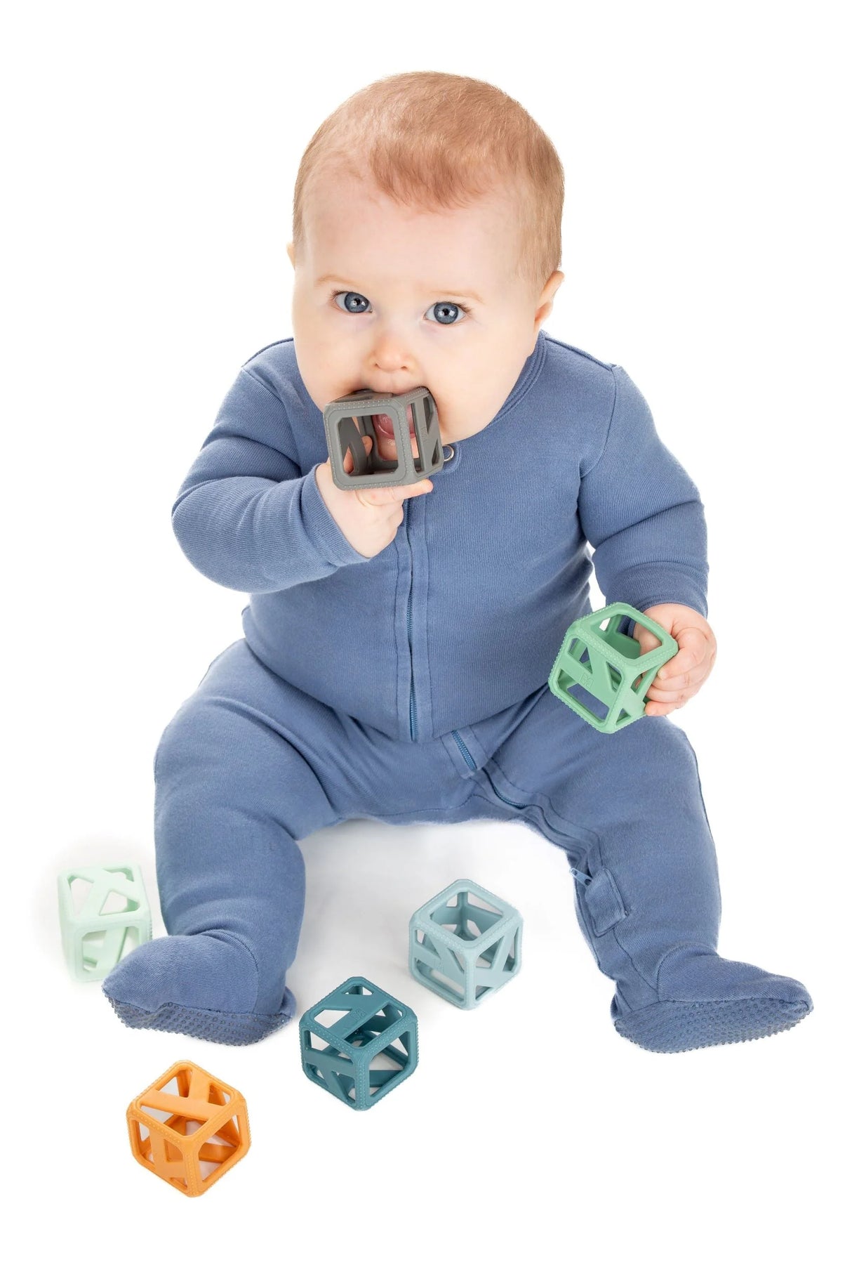 Baby wearing blue onesie chewing and holding Stack & Chew Earthy Cubes teething toy in various colors.