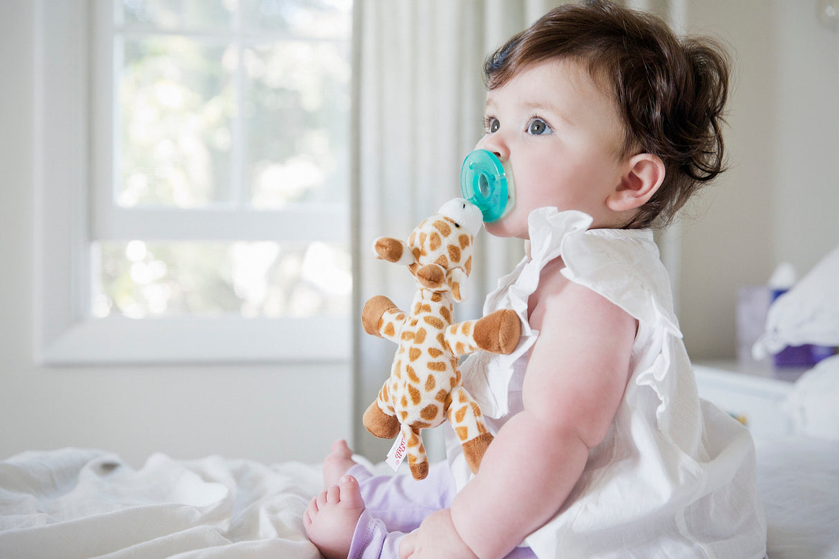 Baby using WubbaNub Baby Giraffe pacifier with soft plush toy attached in bright room.