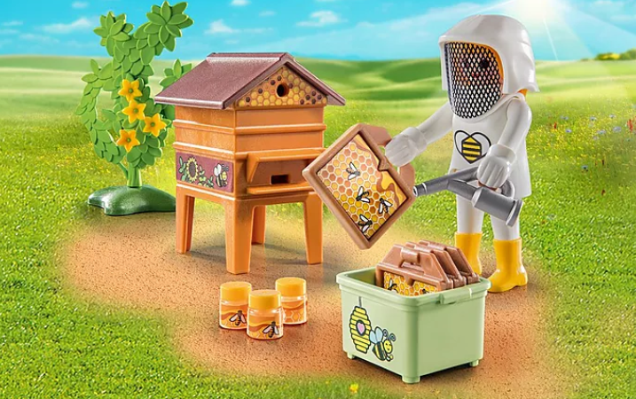 Country | Beekeeper Ali! figure in beekeeping suit harvesting honeycombs with hive, jars, and smoke blower on grass.