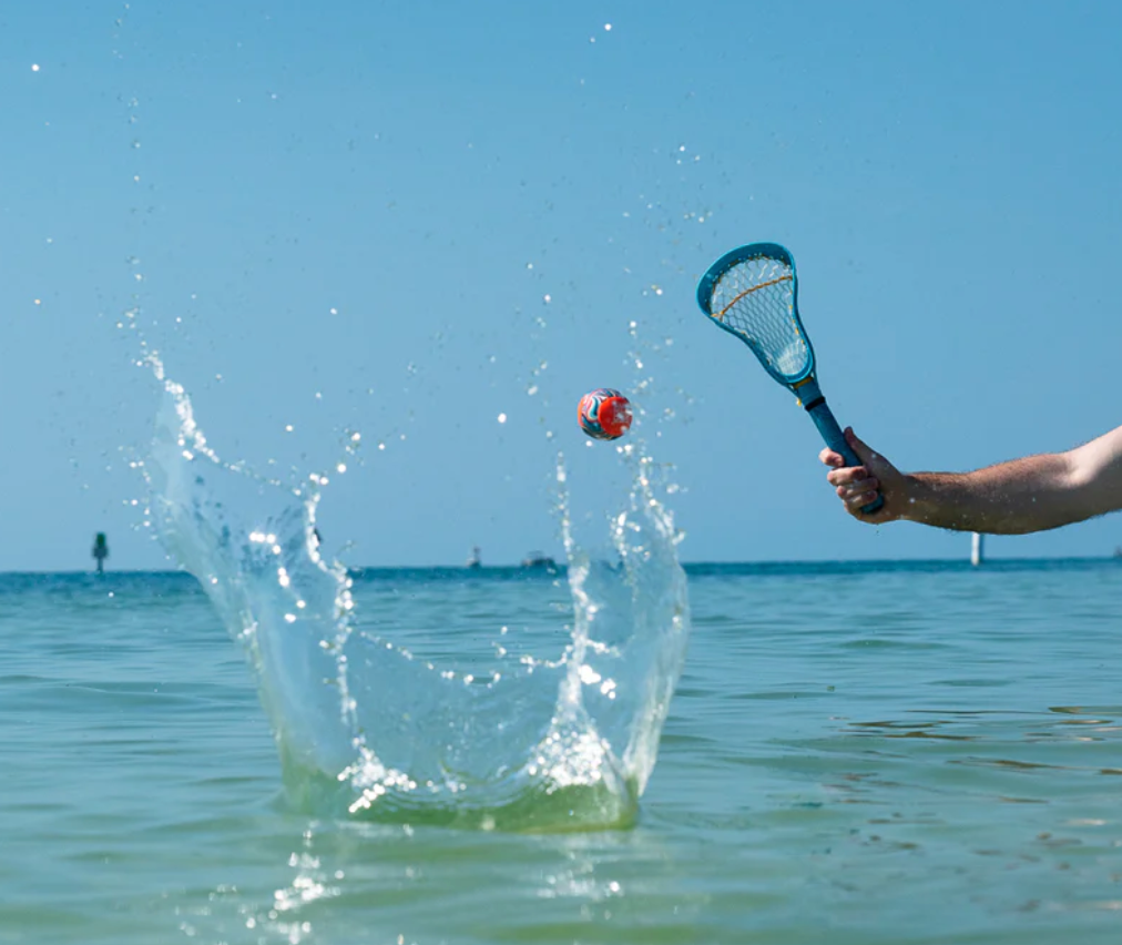 Waboba Mini Lacrosse Set shown in action, with a lacrosse stick and ball bouncing on the water’s surface on a sunny day.
