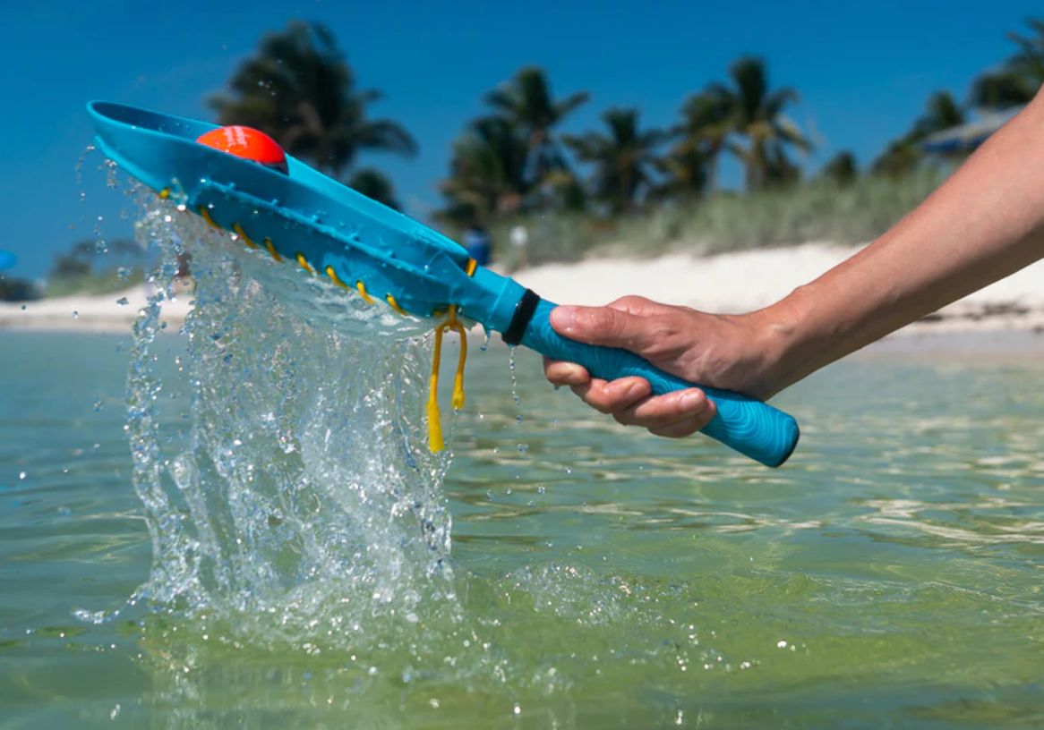 Hand holding Waboba Mini Lacrosse Set stick with orange ball splashing water on a sunny beach day