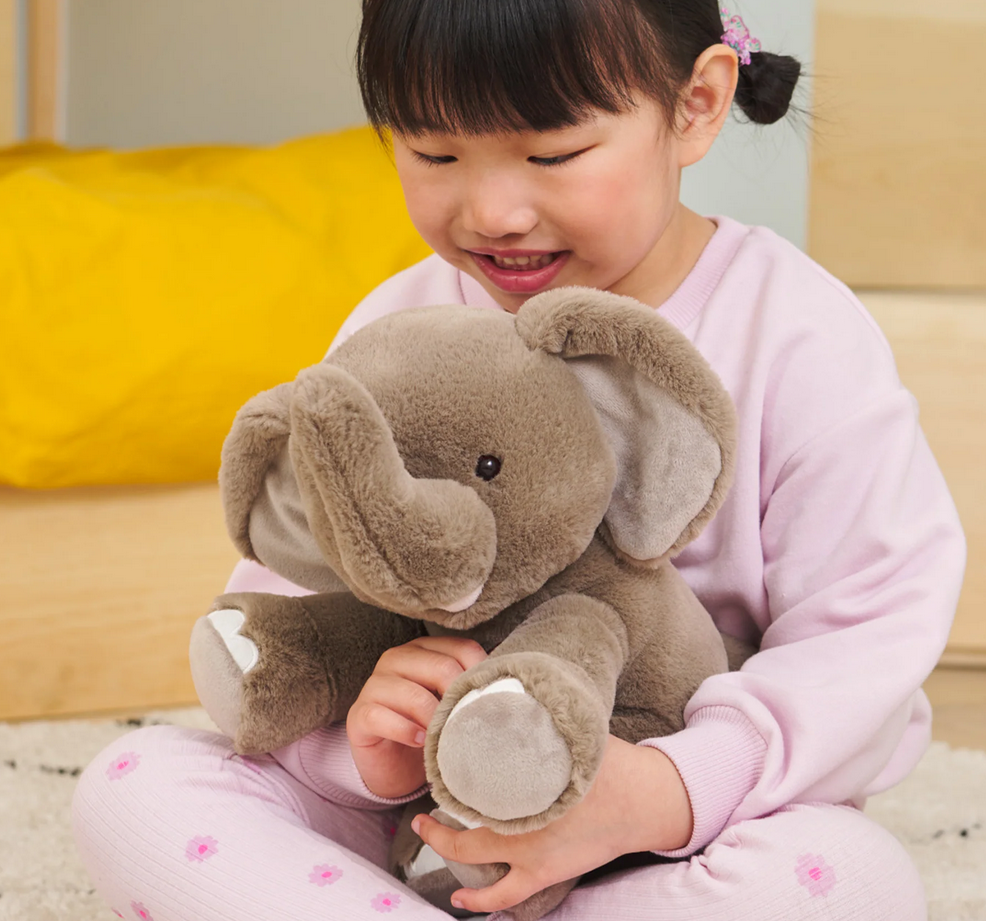 Elephant Chai plush toy in gray soft fabric held by a smiling child sitting indoors on a rug.