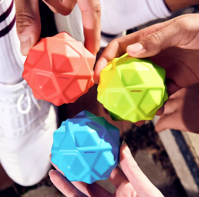 Three hands holding Aerobie Sonic Bounce Ball in red, yellow, and blue with geometric spikes for high bounce.