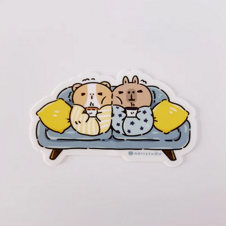 Bubu & Moonch Blanket Burritos Sticker showing cozy guinea pig and capybara wrapped in blankets on a blue couch.