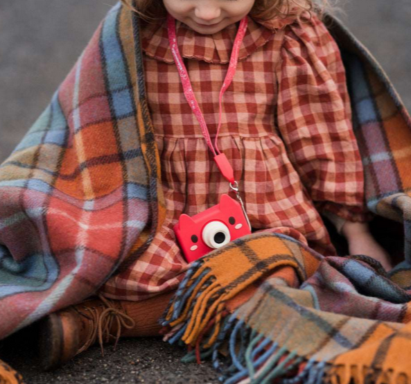 Child wrapped in a colorful plaid blanket holding Digital Camera Model C | Akito the Fox with red silicone case.
