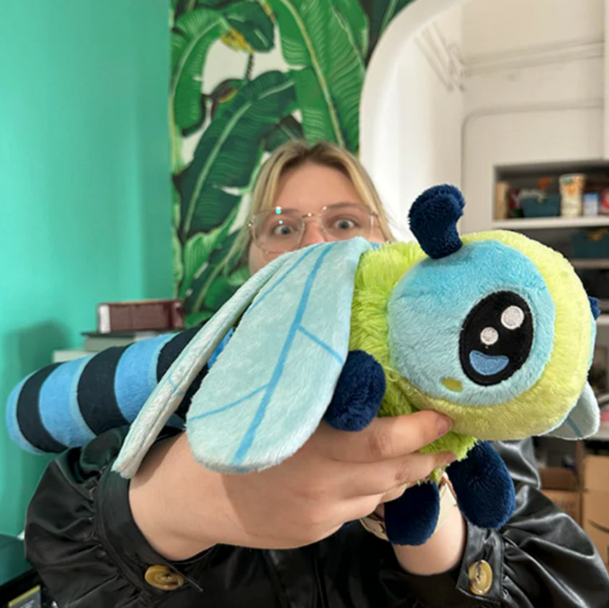 Person holding a colorful plush toy Dragonfly with blue wings and green body indoors.