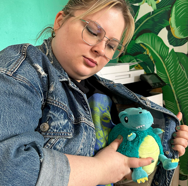 Person holding a soft, blue and green T-Rex Snackers squishable toy inside a denim jacket.