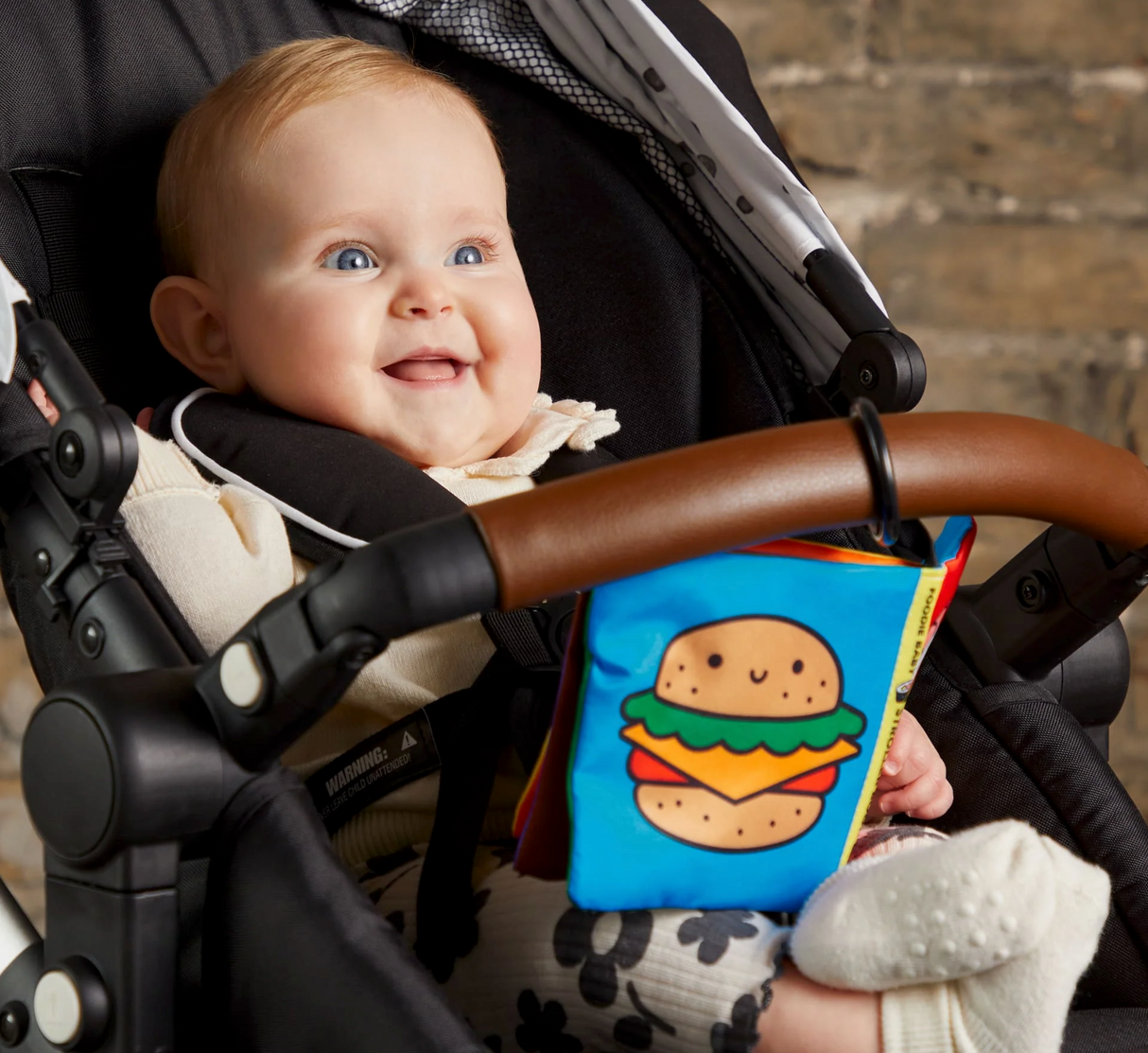 Happy baby sitting in stroller playing with Foodie Baby Crinkle Stroller Book featuring colorful fabric pages.