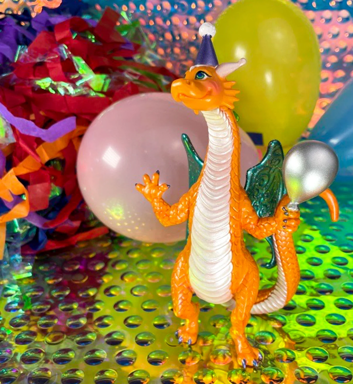 Party Dragon figure wearing party hat and holding balloon, surrounded by colorful balloons and streamers.