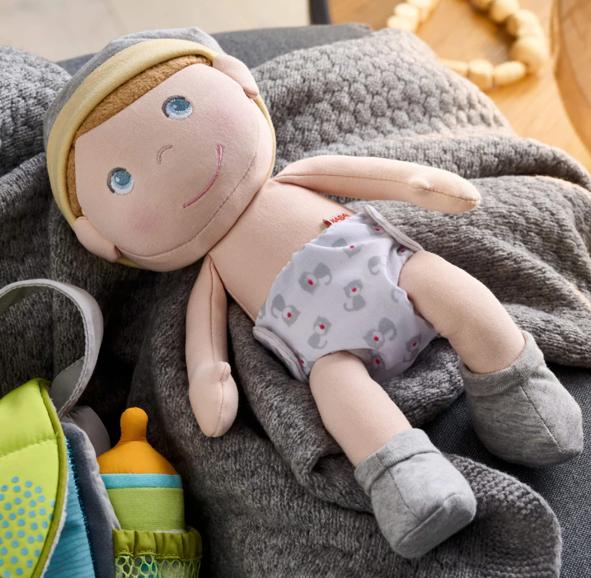 Maxime Baby Doll with embroidered face, reversible diaper, cap, and removable socks lying on a soft blanket.