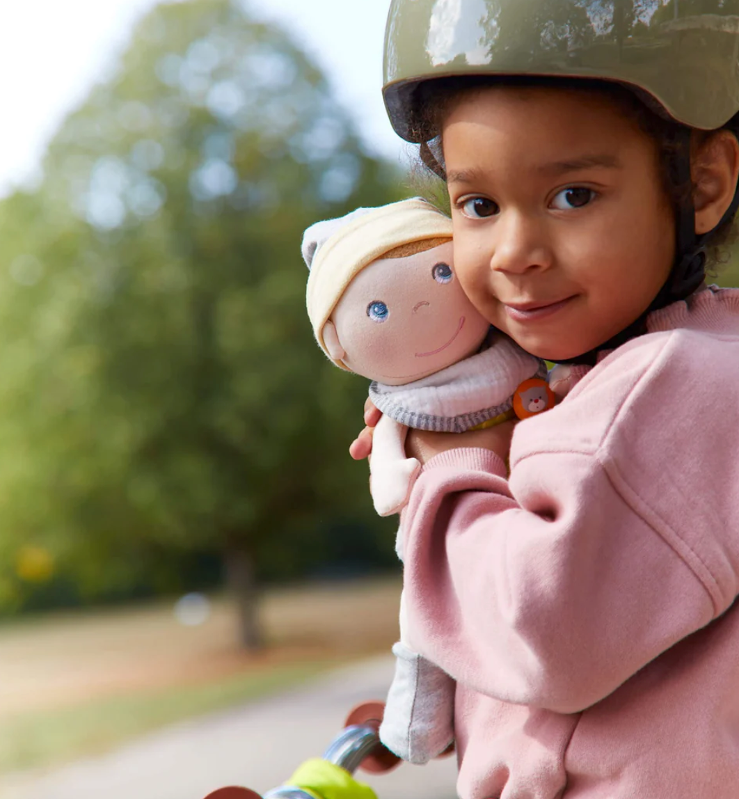 Child wearing helmet hugging Maxime Baby Doll outdoors, showcasing the soft doll with embroidered face and cozy outfit.