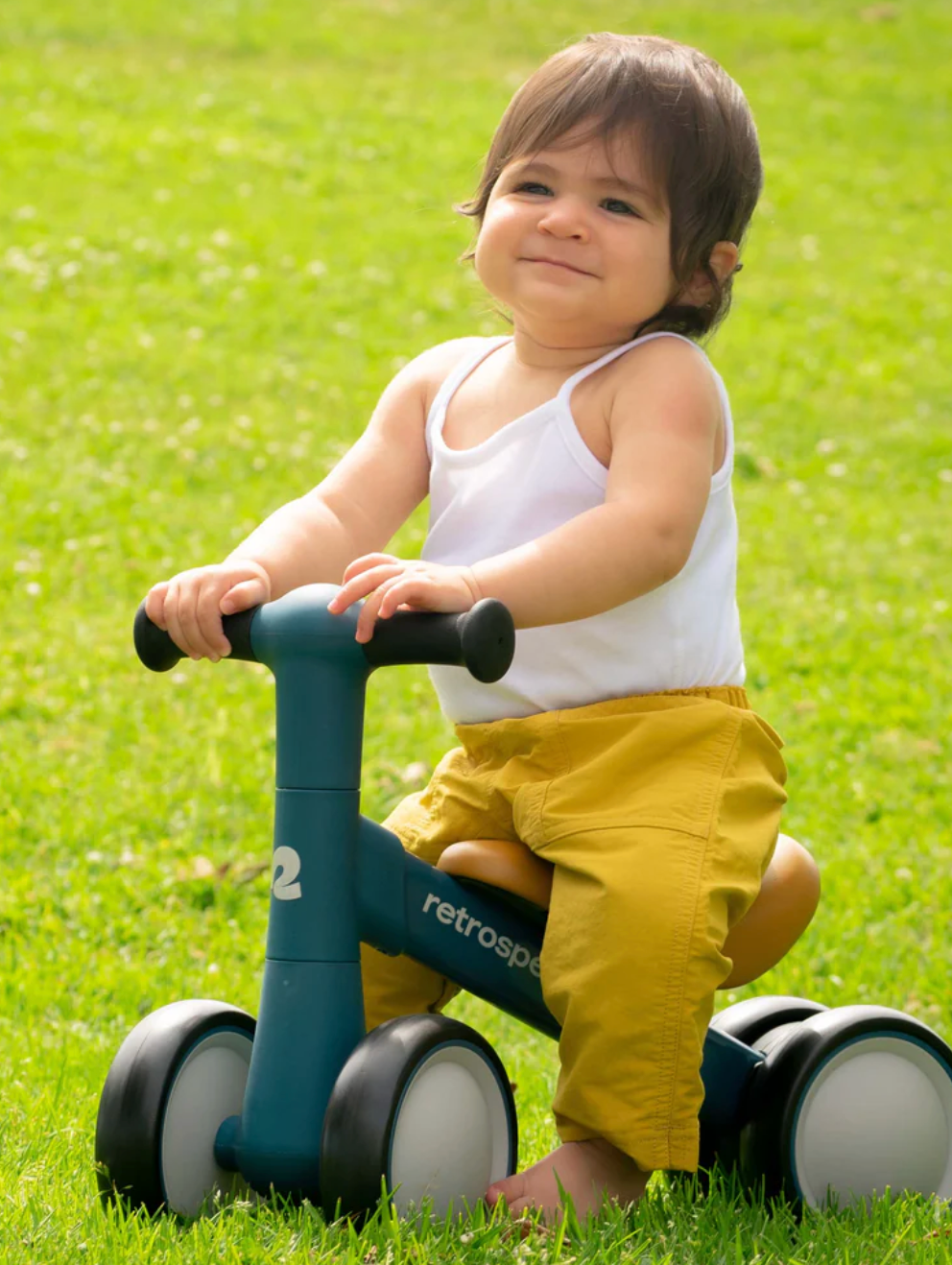 Baby riding a blue Cricket 2 Baby Walker Balance Bike | Baby Pink on grass, developing balance and motor skills.