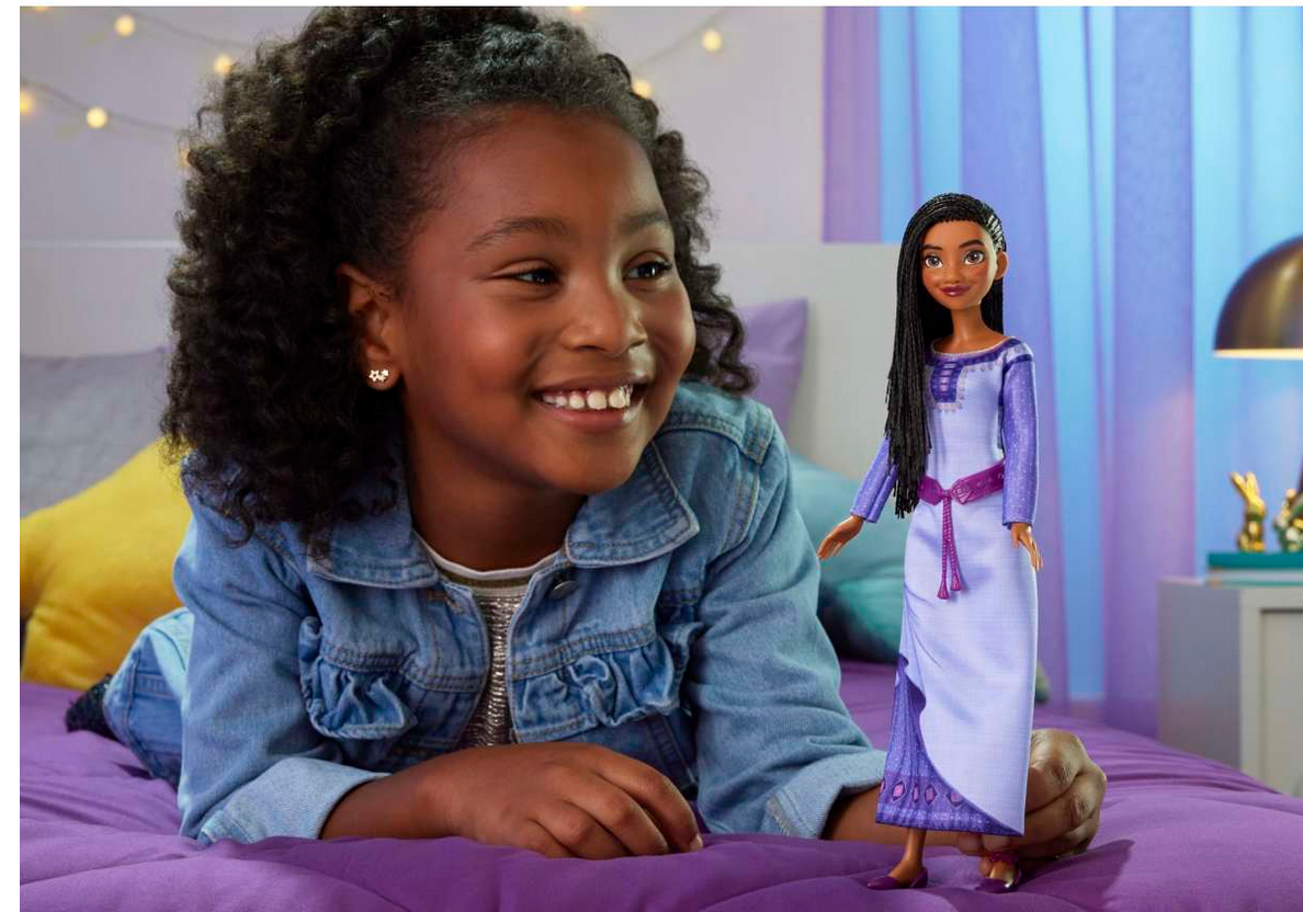 Happy young girl playing with Disney Wish Asha doll in a purple outfit on a bed in a cozy room setting.