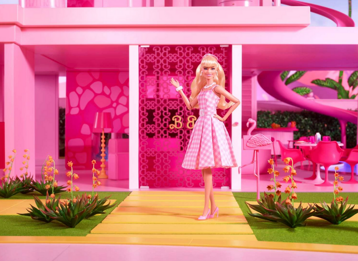 Barbie The Movie doll in pink gingham dress with pastel pumps and daisy necklace outside pink Barbie Land house.