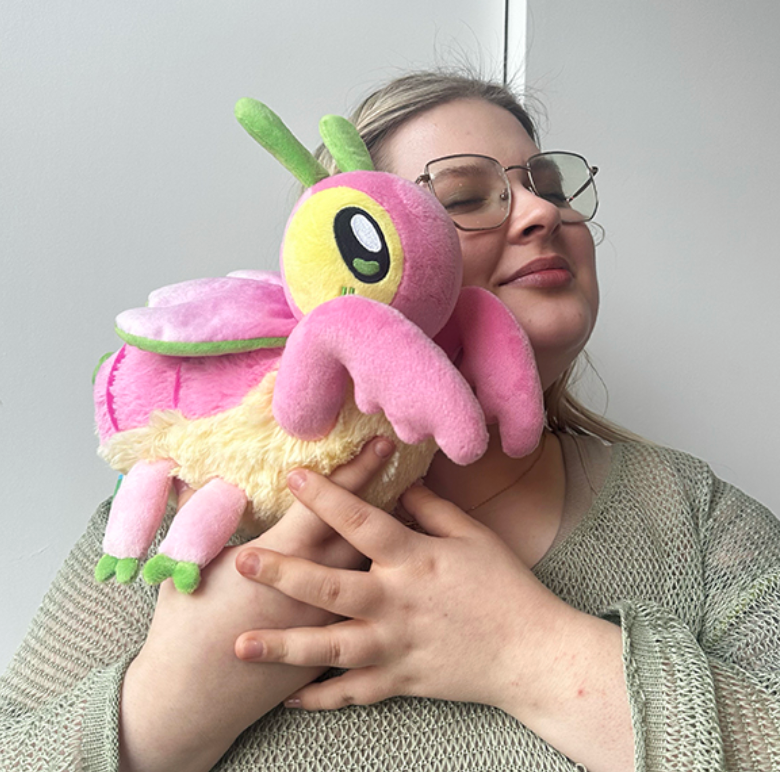A person happily hugging the colorful plush Orchid Mantis Mini with pink wings and green antennae.