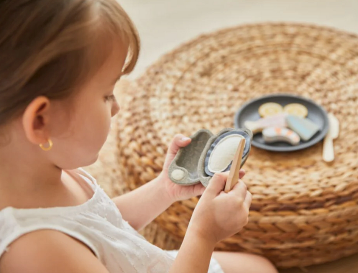 Child playing with PlanToys Seafood Platter, using oyster knife to shuck pretend oyster in a playful kitchen setting.