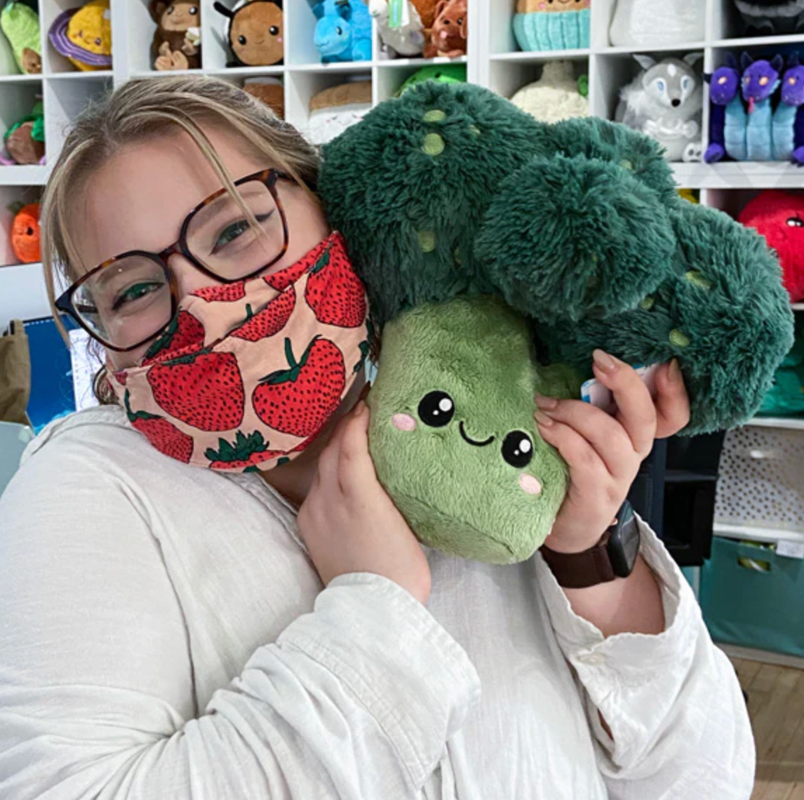 Person wearing glasses and strawberry mask holding a soft plush toy Broccoli Mini with a smiling face.