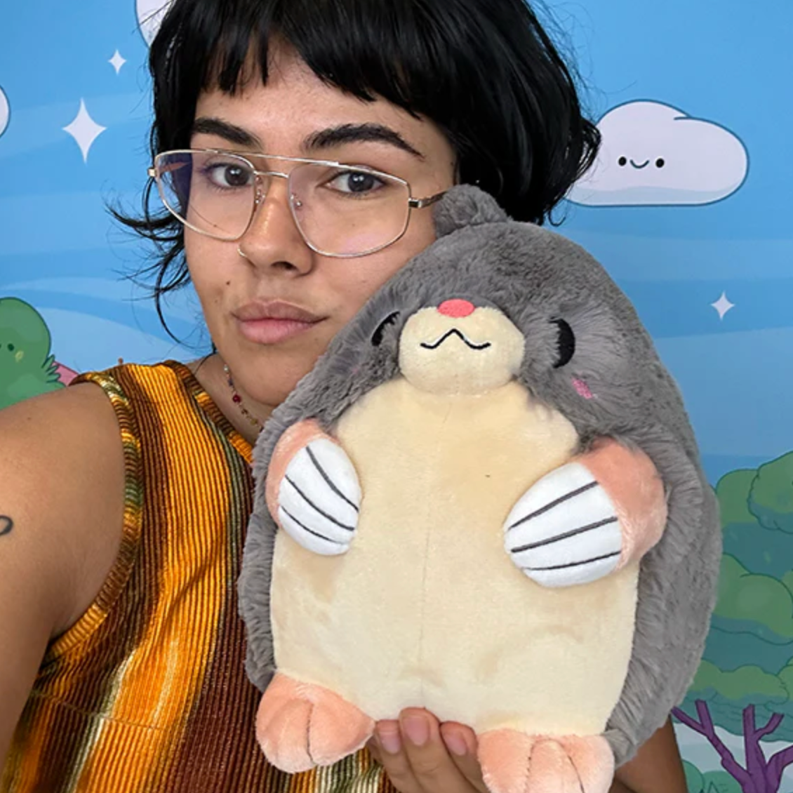 Person holding a soft plush toy Mole Mini with gray fur and pink paws against a colorful backdrop.