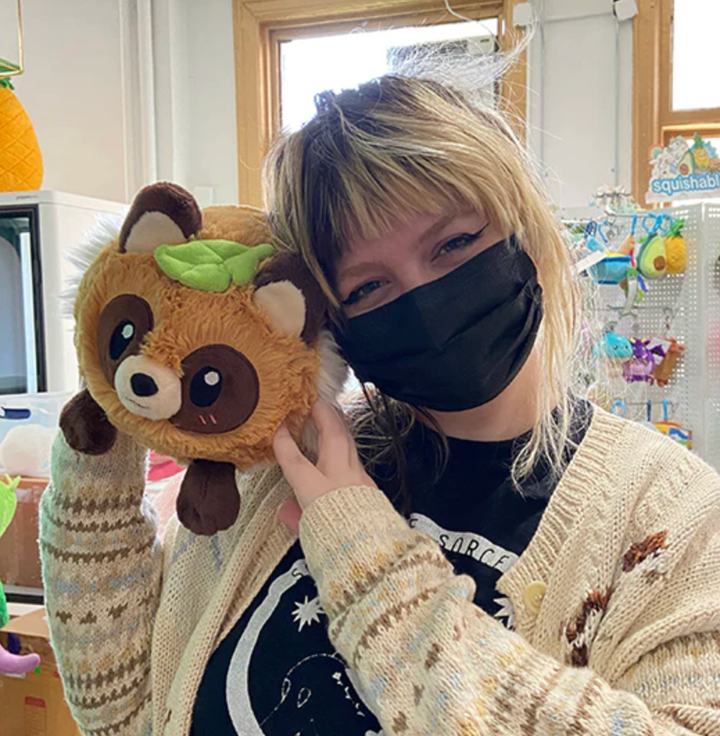 Person wearing a black mask holding a plush Tanuki Mini toy with a green leaf on its head next to their face.