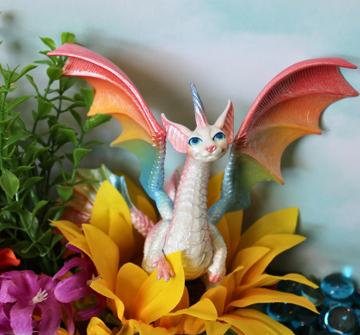 Fairy Rainbow Dragon figurine with pearlescent body and colorful rainbow wings sitting among bright flowers.