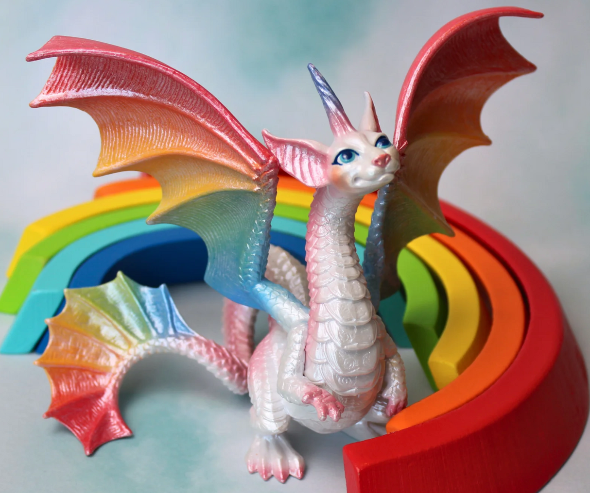 Fairy Rainbow Dragon figurine with pearlescent body and vibrant rainbow wings posed in front of colorful rainbow arches.