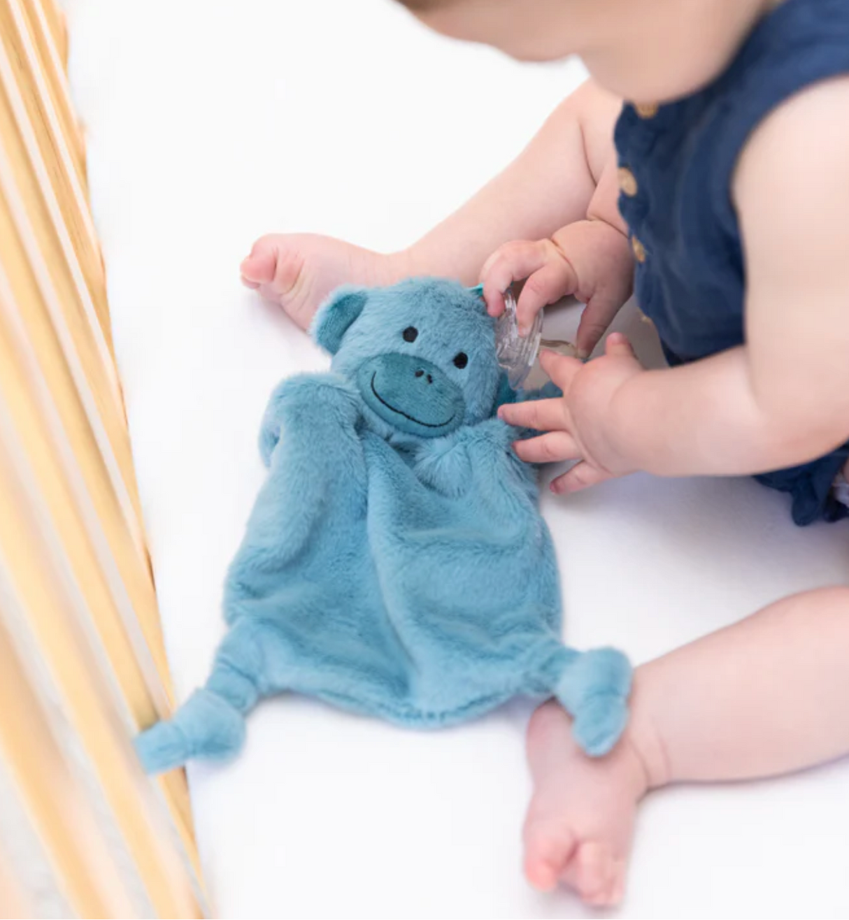 Baby playing with blue plush Milo the Monkey pacifier buddy with pacifier loop on white crib mattress.