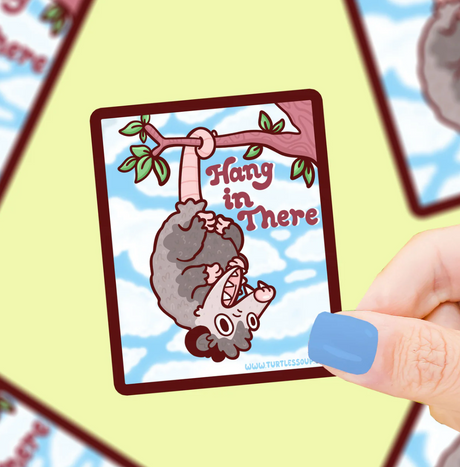 Hand holding a Hang In There Opossum Sticker featuring a cute possum hanging from a tree branch with blue sky background.