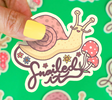 Hand holding Snailed It Sticker featuring a happy snail with flowers and mushrooms on a bright green background.