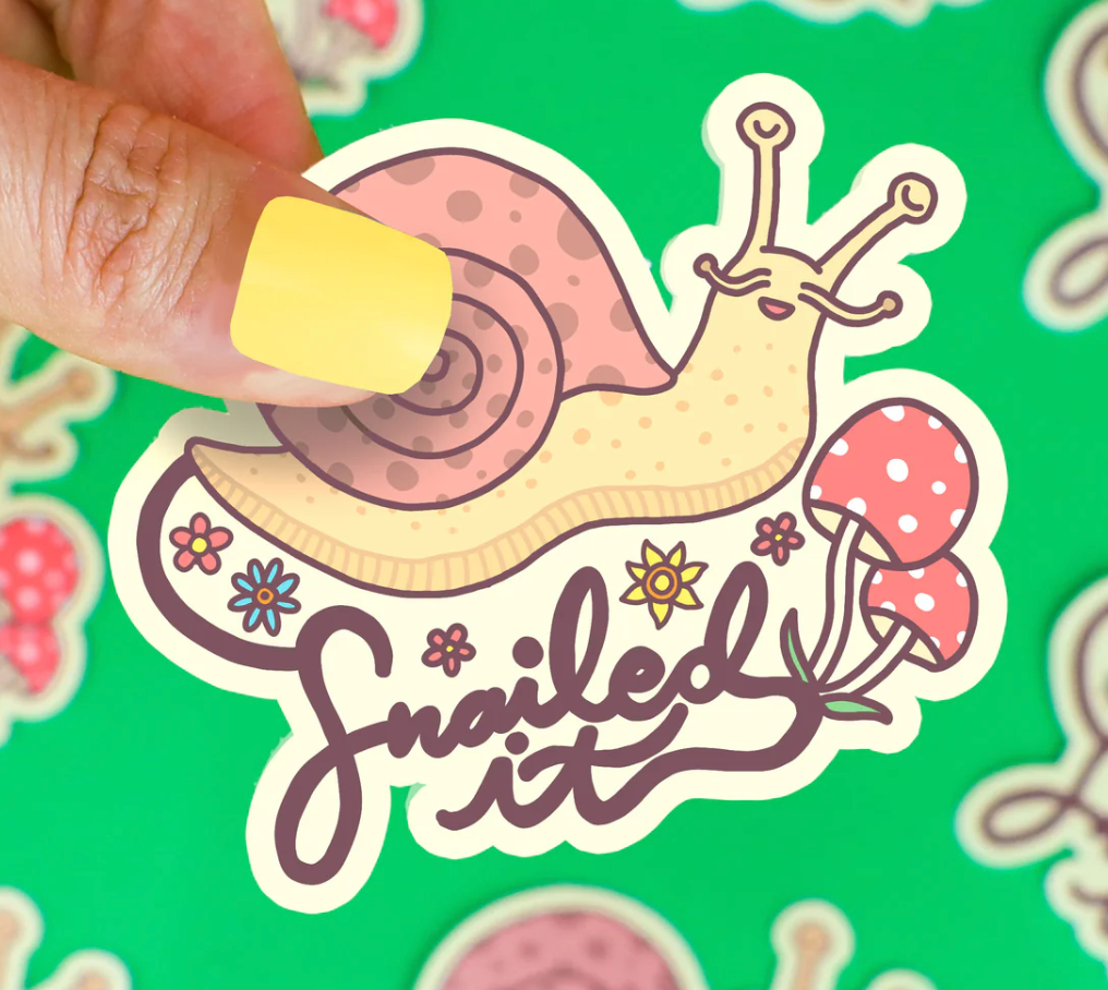 Hand holding Snailed It Sticker featuring a happy snail with flowers and mushrooms on a bright green background.