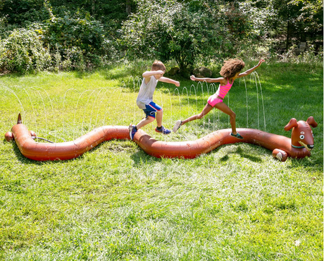 Kids jumping over the Sprinkler | Long Dog inflatable wiener dog sprinkler spraying water in a sunny backyard.