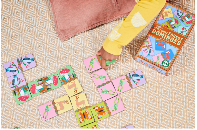 Child playing with Giant Shiny Forest Dominoes featuring colorful forest creatures on a woven rug beside the game box.