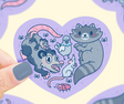 Hand holding Stinky Trash Animals Heart Sticker featuring cute trash animals inside a purple heart shape.