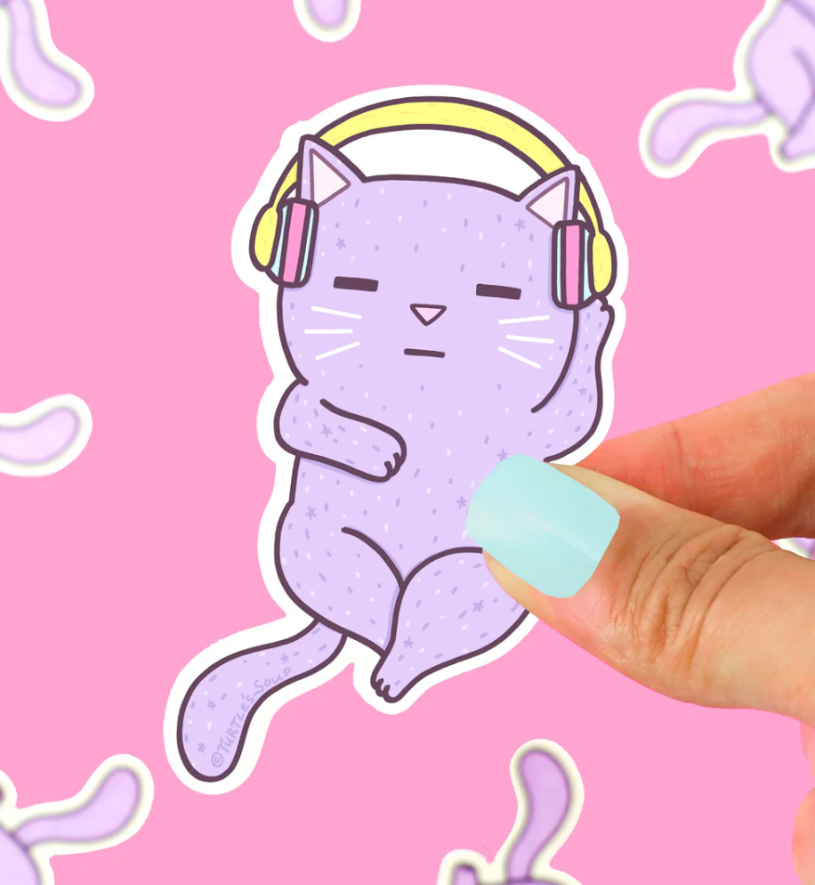 Hand holding a purple cat sticker wearing headphones, titled Headphones Cat Sticker, against a pink background.