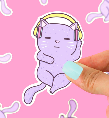 Hand holding a purple cat sticker wearing headphones, titled Headphones Cat Sticker, against a pink background.