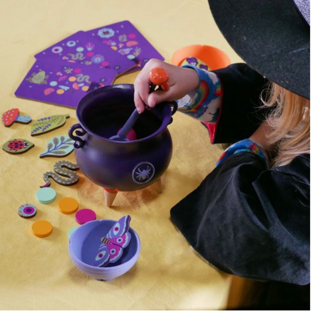 Child in witch costume stirring wooden pieces in a purple cauldron from the Witch's Brew play set.