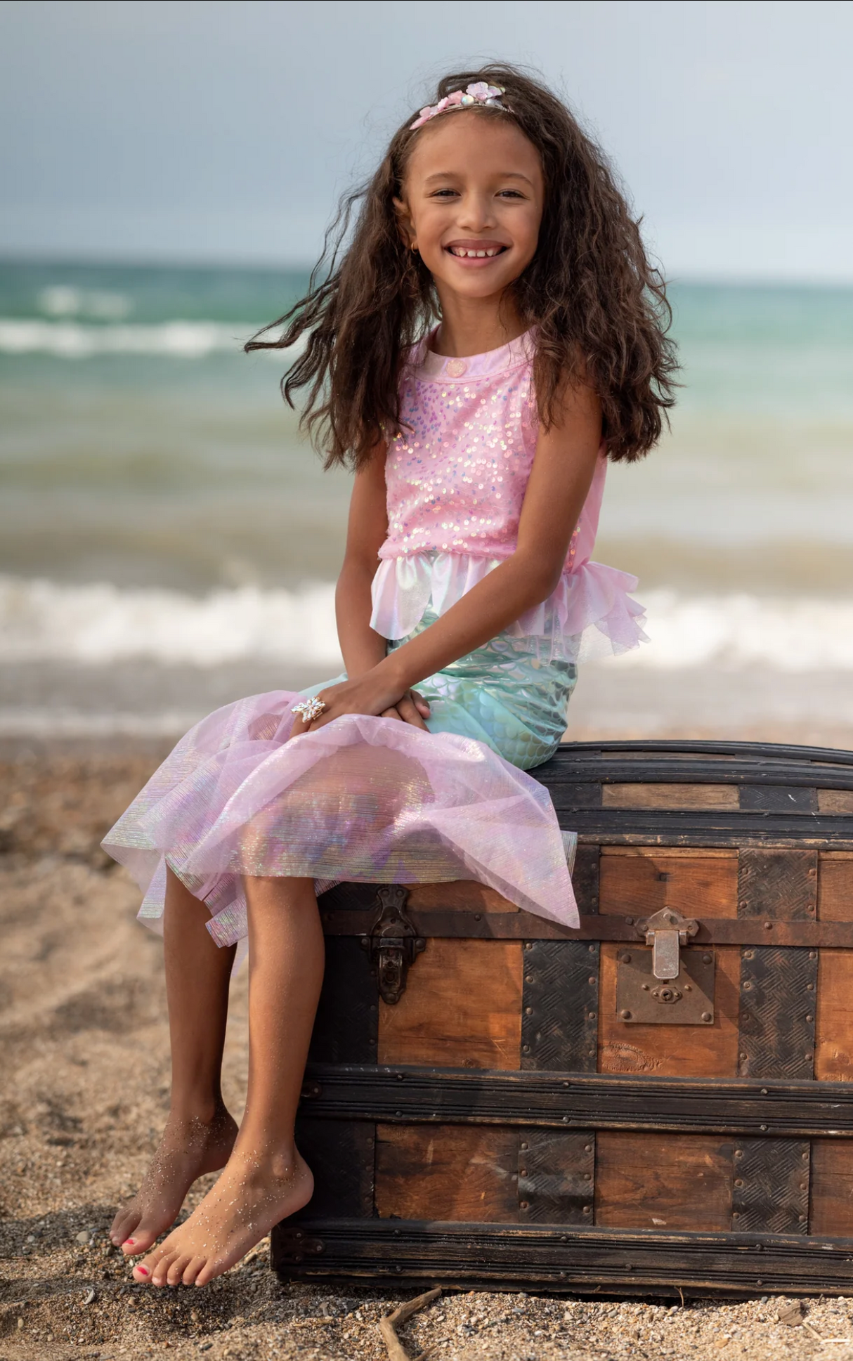 Girl wearing Misty Mermaid Dress Pink with sequins and tulle skirt sitting on a wooden chest by the beach.