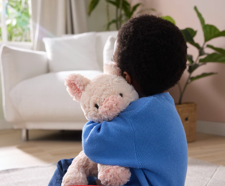 Child hugging a soft, textured Cozys Pig plush toy with peachy-pink fur and floppy limbs for cuddling.