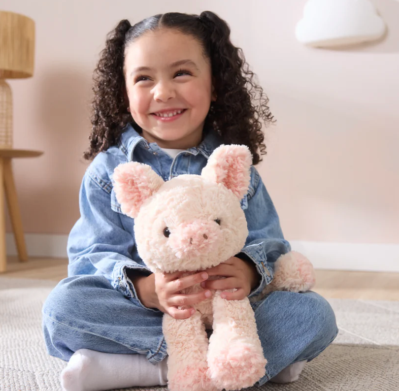 A smiling child cuddling a soft, textured Cozys Pig plush toy with peachy-pink fur and floppy limbs.