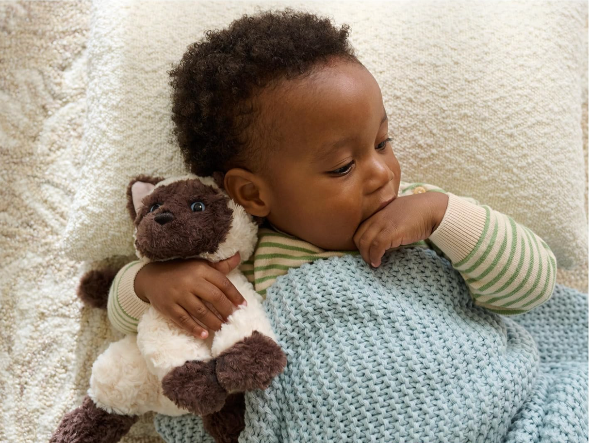 Child cuddling a Bootsie Siamese Kitten plush toy with cream and chestnut fur, lying under a blue blanket.