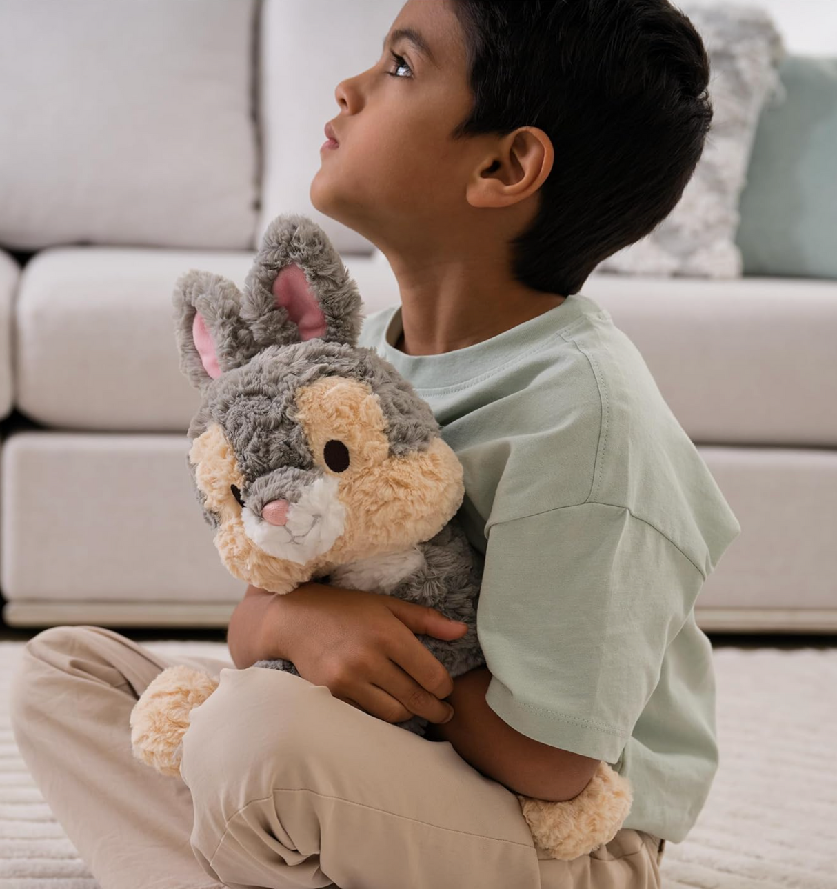 Young boy sitting on floor hugging a soft, gray and cream Cozys Thumper plush bunny with pink ears.
