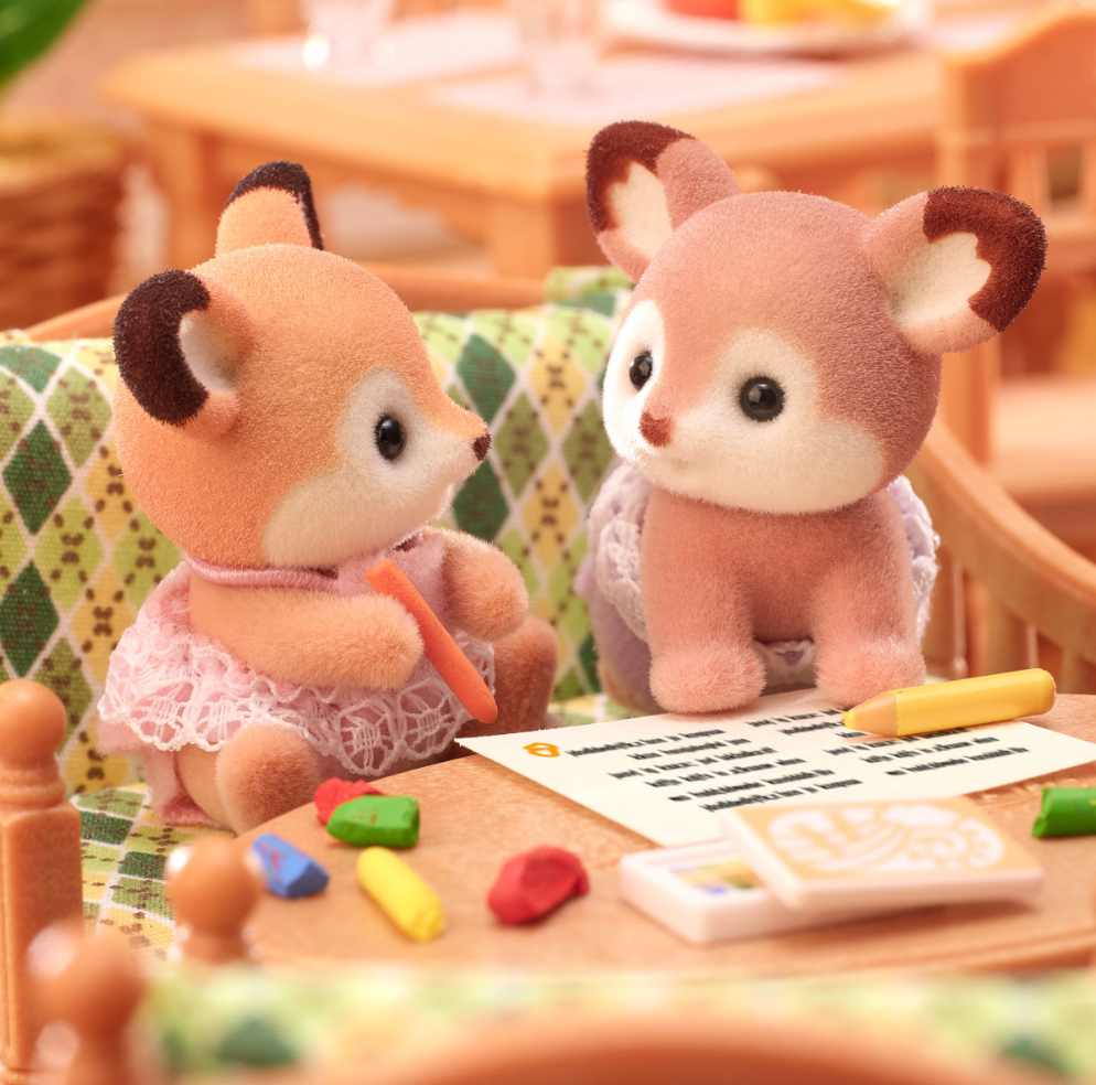 Deer Twins plush figures with big ears sitting on a patterned couch surrounded by crayons and paper.