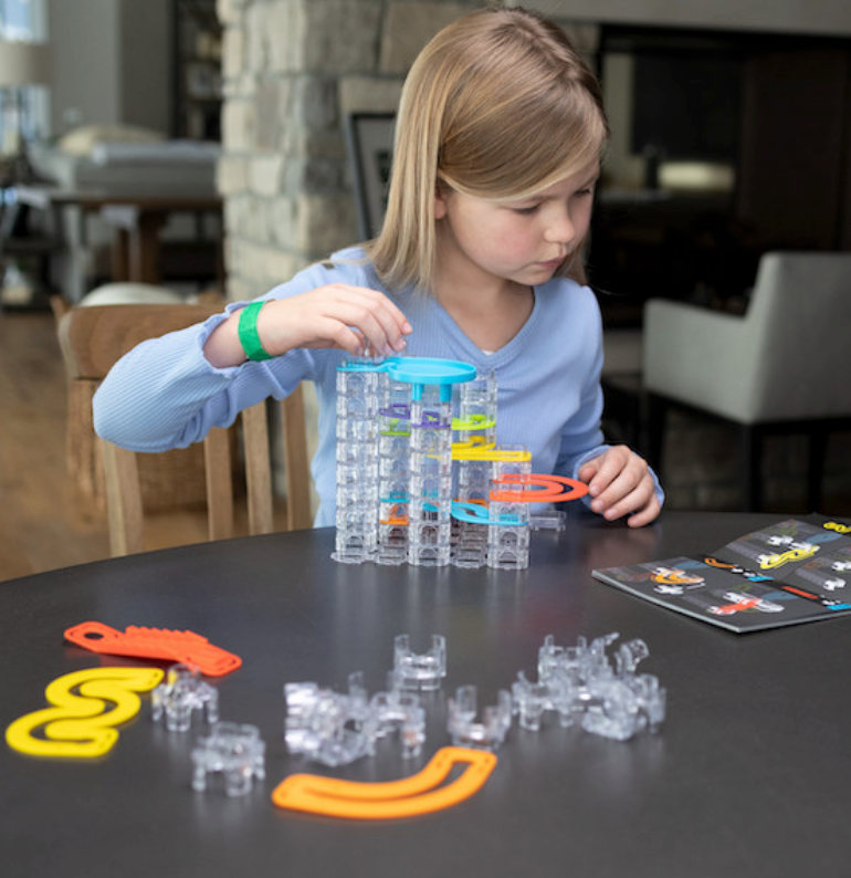 Young girl assembling the Trestle Tracks Builder Set with clear stacking cubes and colorful tracks on a table.