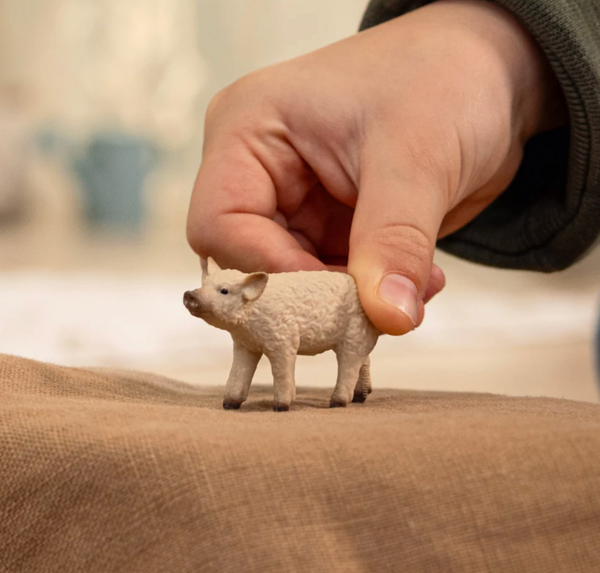 Hand holding a small beige curly-coated Mangalica Piglet toy with black eyes on a brown surface.