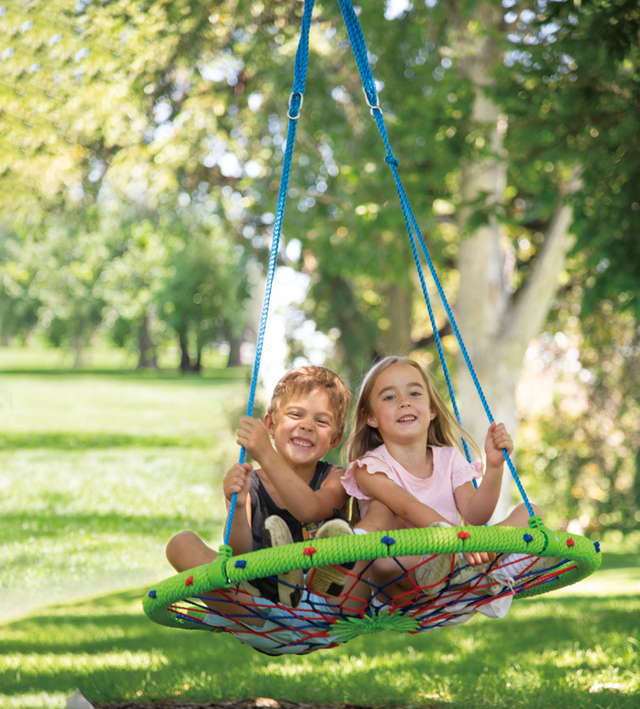 Two children enjoying playtime on a colorful Dreamcatcher Swing hanging from tree branches outdoors.