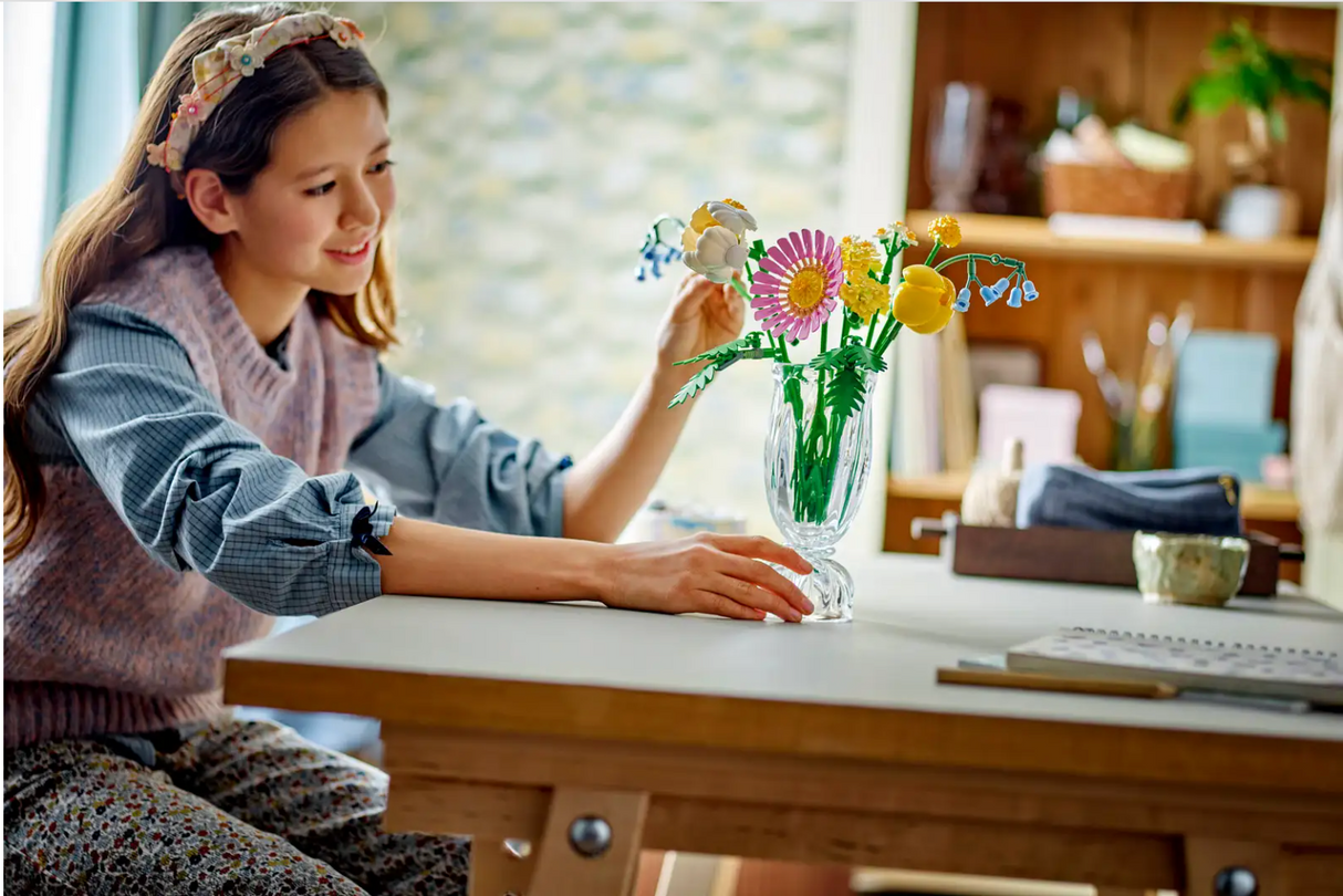 A girl arranges a colorful Botanicals Petite Sunny Bouquet of LEGO spring flowers in a clear vase on a wooden table.