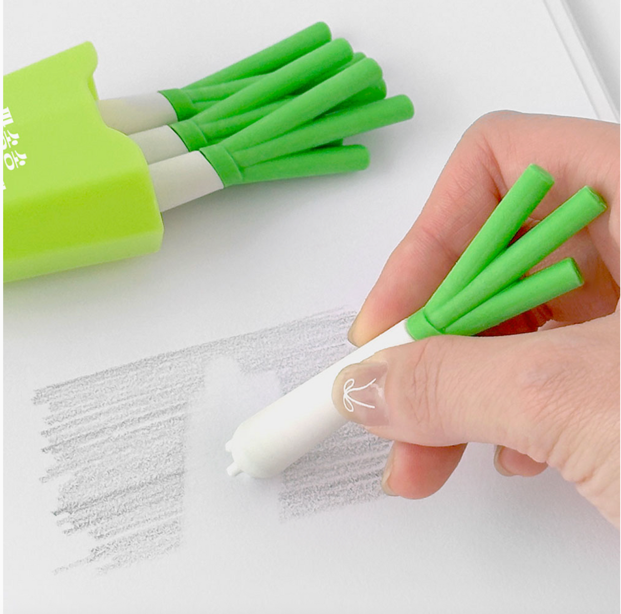 Hand holding a green onion shaped eraser erasing pencil marks with more erasers in a green case nearby